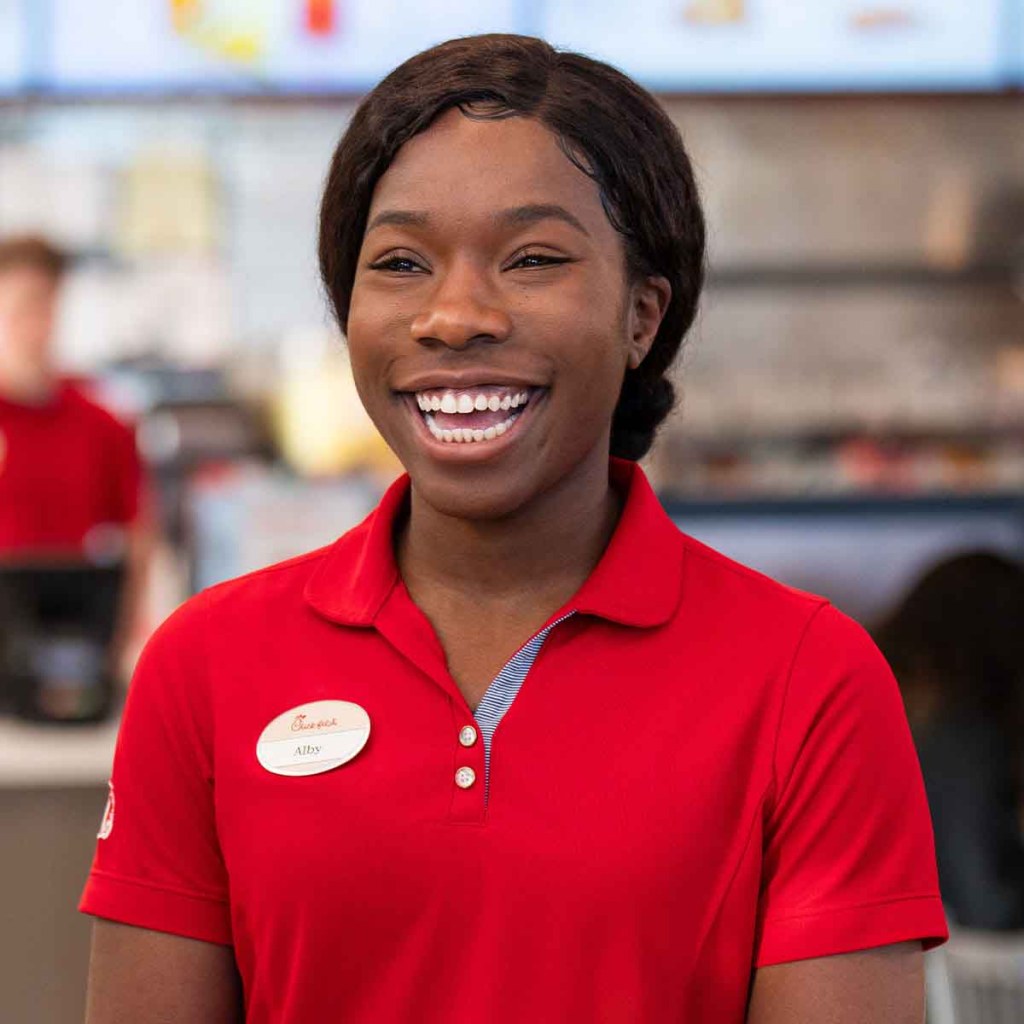 Smiling Chick-fil-A team member Alby (red polo, nametag, dark hair pulled back) laughs. Blurred restaurant in the background