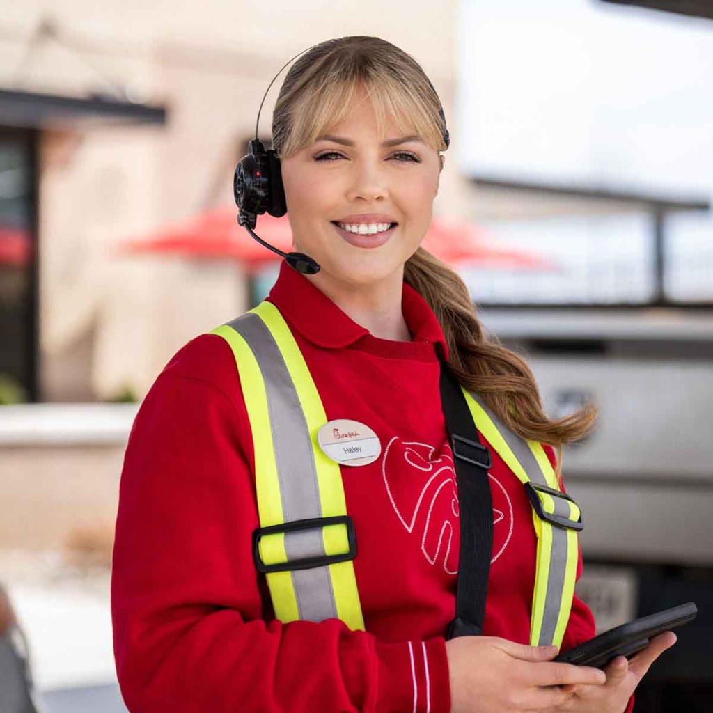 Smiling Chick-fil-A team member Haley (blonde hair, red long-sleeved shirt, nametag, headset, hi-vis vest) looks at camera. 