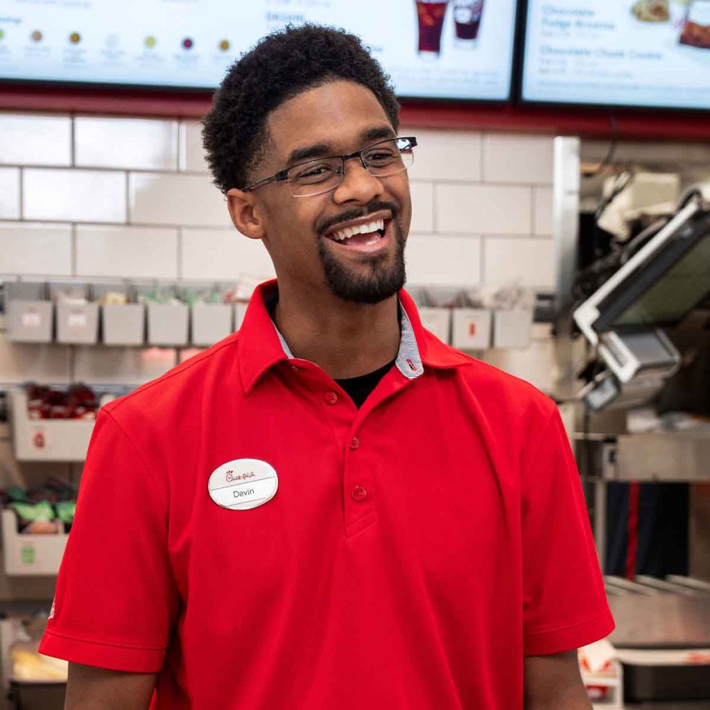 Smiling Chick-fil-A team member (man, curly hair, beard, glasses, red polo) laughing, looking right. Menu boards in the background 