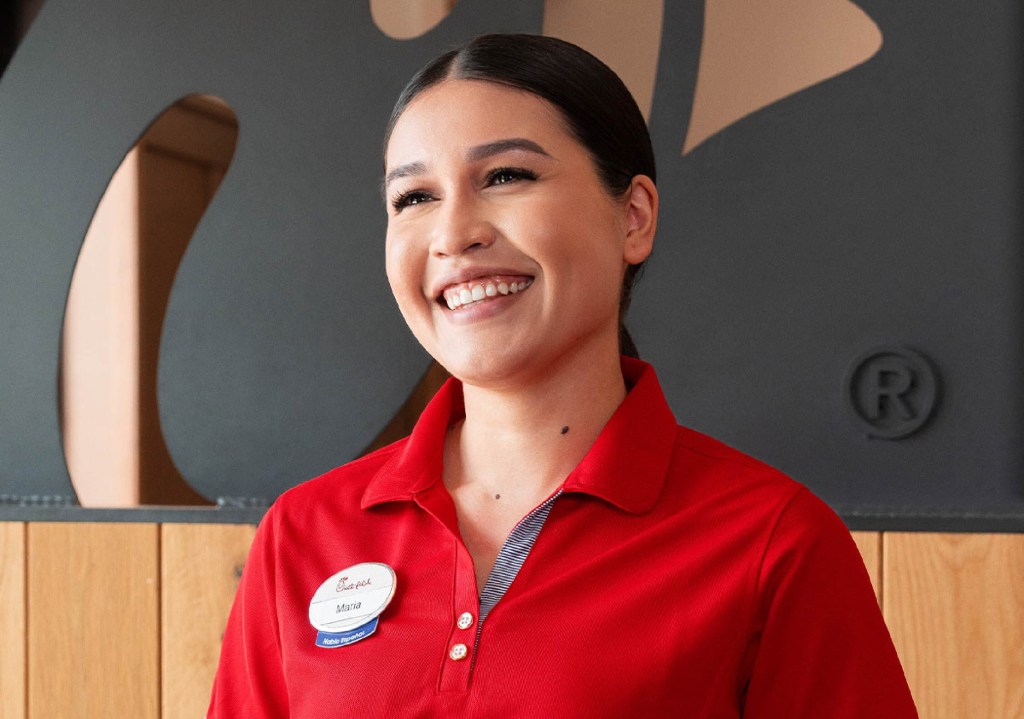 Smiling Chick-fil-A team member Maria (dark hair, red polo, nametag) looks right. Chick-fil-A logo in background.
