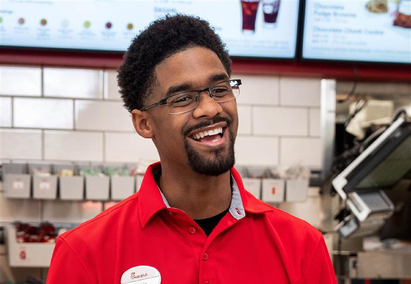 Smiling Chick-fil-A team member (man, curly hair, beard, glasses, red polo) laughing, looking right. Menu boards in the background