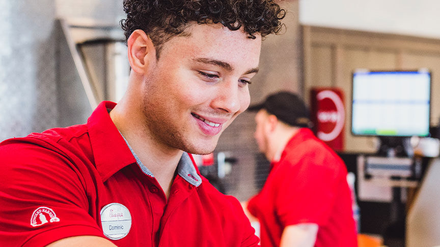Smiling Chick-fil-A team member Dominic (curly brown hair, red polo) with a "Scholarship Recipient" patch on his sleeve